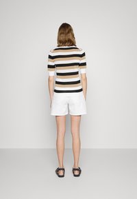 Kvinde står med ryggen til iført en beige, sort og hvid stribet skjorte, hvide shorts og sorte sandaler på en grå gulvbaggrund.