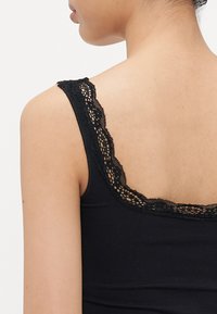 Femme vue de dos, avec le haut du dos et l'épaule visibles, portant un top sans manches noir agrémenté de dentelle au niveau de l'encolure et de la bretelle.
