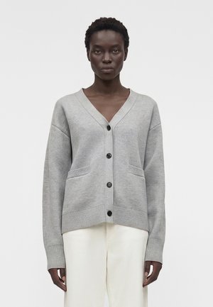 Žena stojaca, oblečená v svetlosivej zapínacej kardigáne s prednými vreckami a biele nohavice na jednotnom bielom pozadí.