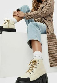 Klara platformskor i krämfärg med svarta sulor, prydda med metalliska öljetter och guldfärgade skosnören. Bärs med ljusblå korta jeans och vita strumpor.