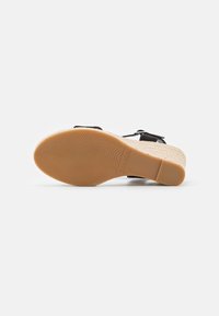 Sandal con tacco a cuneo e suola in gomma texturizzata, con cinturini neri e piattaforma intrecciata beige, mostrata dal basso su uno sfondo bianco.