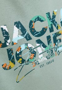 Tissu vert clair avec un imprimé graphique audacieux présentant "JACK & JONES ESSENTIALS" dans un style éclaboussé multicolore, avec l'année d'établissement 1990.
