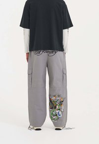 Graue Cargo-Hosen mit bestickten Designs, die einen Schädel und einen Adler zeigen, verstellbare Bündchen und Ed Hardy-Branding am Bund. Schwarzes T-Shirt darunter geschichtet.