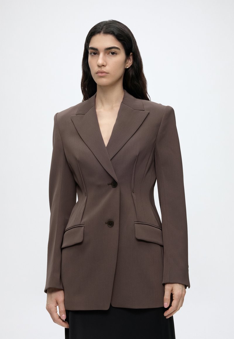 Jeune femme aux longs cheveux foncés porte un blazer marron sur mesure avec deux boutons et des poches à rabat, debout devant un fond uni.
