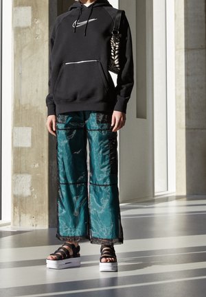 Personne portant un sweat à capuche Nike noir, un pantalon large bleu sarcelle avec des patches noirs, des sandales à semelles compensées noires et un sac à bandoulière à rayures zébrées.