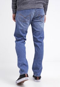 Jean en denim bleu avec une coupe décontractée, présentant des poches arrière avec un motif de couture subtil, portés avec des baskets noires.