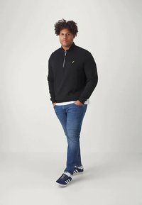 Black zip-up sweatshirt, white T-shirt underneath, blue jeans, and blue sneakers. Model stands with hands in pockets against a light background.