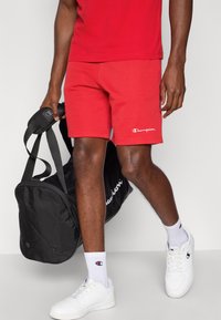 Κόκκινα αθλητικά σορτς από βαμβάκι με λευκό λογότυπο, συνδυασμένα με λευκά αθλητικά παπούτσια και γκρι κάλτσες έως τον αστράγαλο, κρατώντας μια μαύρη αθλητική τσάντα.