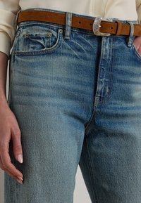 Pogled na modre denim hlače z blaznimi podrobnostmi in rjavim semiš pasom z okrasno srebrno sponko, nosijo pa se z smetanasto belim zgornjim delom.