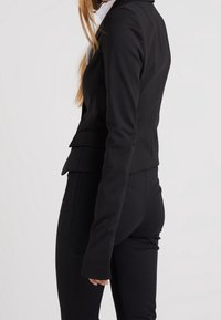 Svart skräddarsydd kavaj med en strukturerad silhuett, slätt tyg och långa ärmar, samt en framträdande slagkrage och diskreta veck vid midjan.