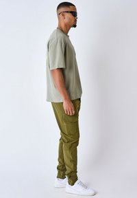 Pantalones cargo verdes oliva con bolsillos laterales, combinados con una camiseta verde clara. Zapatillas blancas completan el atuendo. Incluye un elegante collar de cadena.