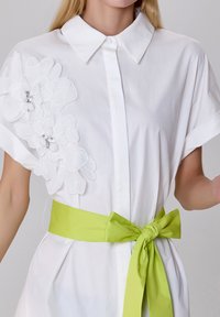 Robe blanche à manches courtes, ornée d'une broderie florale sur le côté gauche et d'une ceinture verte vif nouée à la taille.