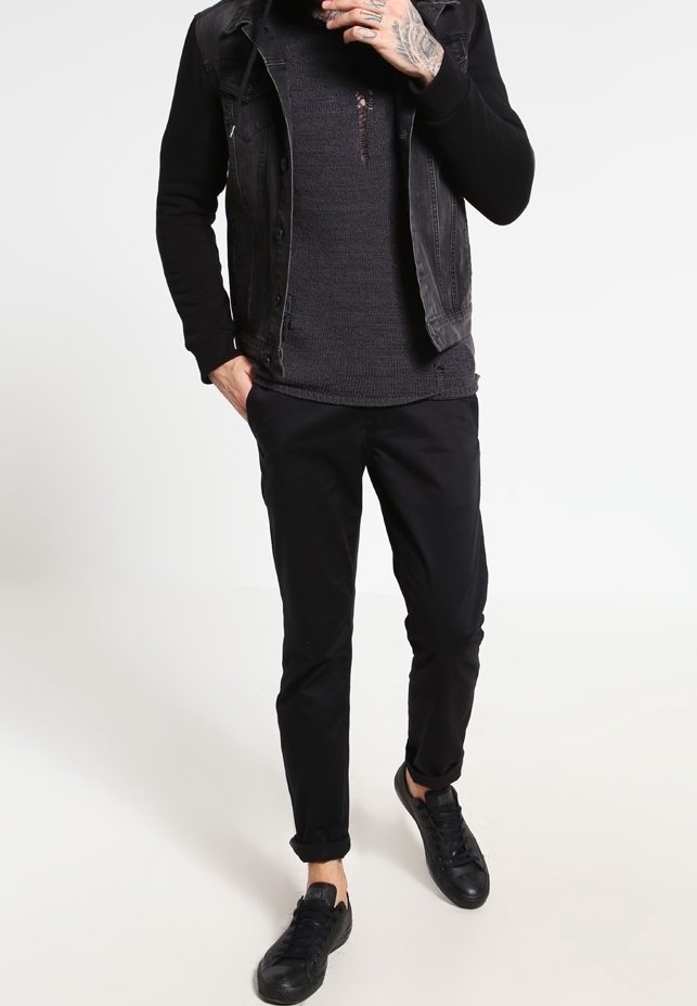 Homme portant un gilet en denim noir, une chemise noire à manches longues, un pantalon noir et des baskets noires, debout avec une main dans la poche.
