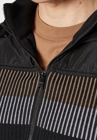 Gros plan d'une personne portant un t-shirt marron à col rond sous un pull zippé noir avec des rayures verticales marron et blanches.