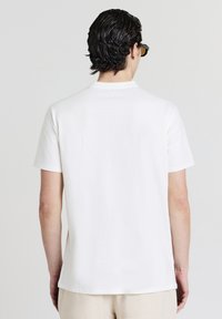 Uomo con capelli scuri e mossi che indossa occhiali neri, una semplice camicia bianca a maniche corte e pantaloni beige chiari, di spalle rispetto alla fotocamera.