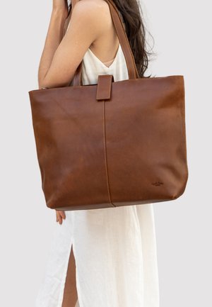 Brown leather tote bag with a smooth texture, spacious design, accented by a top flap closure, displayed against a neutral background.