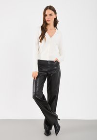 Biely pletený kardigán s výstrihom do V a gombíkmi, skombinovaný s čiernymi koženými nohavicami s širokými nohavicami a čiernymi špicatými podpätkami. Hladké textúry, minimálny dizajn.