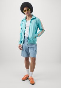 Lyseblå hættejakke med orange striber, hvid t-shirt, denimshorts og orange sneakers. Modellen har afrofrisure og gule solbriller.