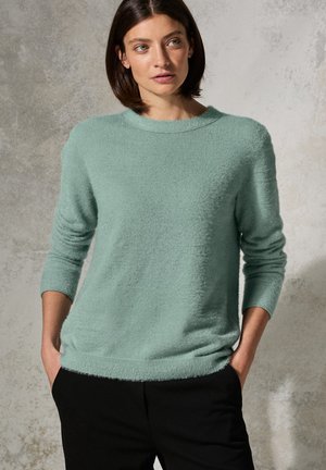 Pull vert clair avec une texture douce, encolure ronde et manches trois quarts, associé à un pantalon noir à coupe décontractée.