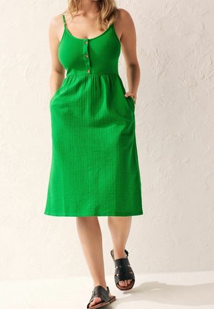 Kobieta w zielonej, bezrękawnikowej sukience midi z guzikami i kieszeniami, w czarnych sandałach z odkrytymi palcami, stojąca na tle fakturowanej ściany.