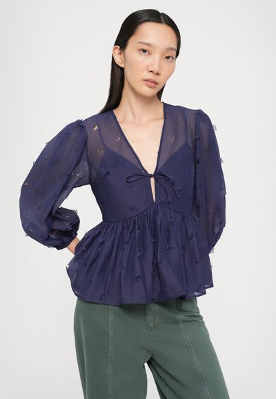 Blusa azul marino, transparente, con escote en V y detalle de lazo. Presenta mangas abullonadas y formas bordadas a lo largo de la tela. Diseño acampanado en la cintura.