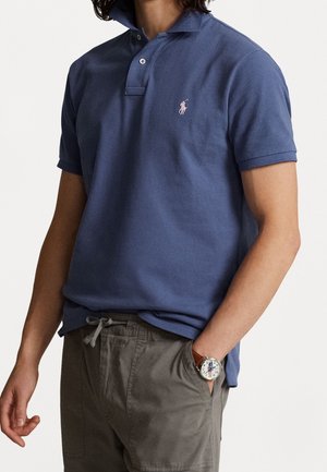 Mann trägt ein blaues Polohemd mit kurzen Ärmeln und Logo, graue Hose mit Kordelzug und eine Armbanduhr mit braunem Lederarmband.