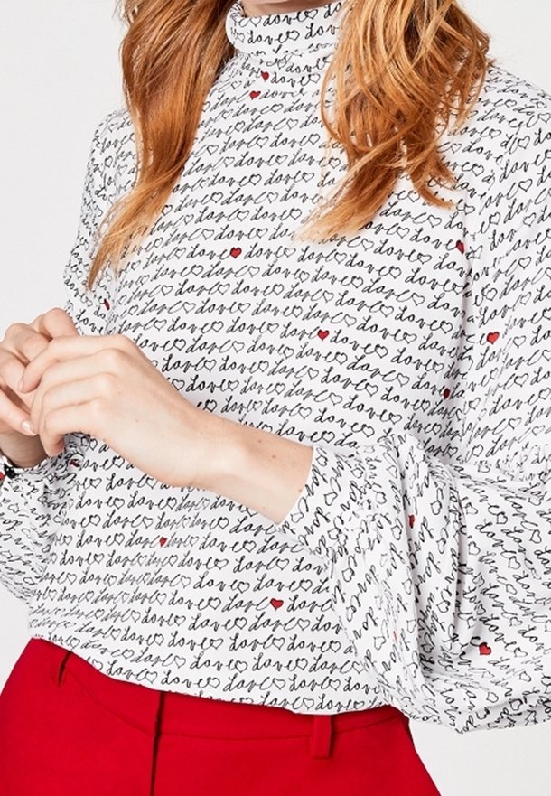 Biała bluzka z długim rękawem z czarnym ręcznie napisanym tekstem i czerwonymi akcentami w kształcie serca; ma wysoki kołnierz i obszerne rękawy.