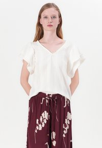 Blusa bianca a maniche corte con scollo a v, caratterizzata da tessuto testurizzato e righe verticali. Abbinata a pantaloni borgogna decorati con motivi floreali.