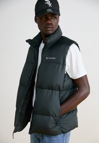 Sort puffervest med høj krave, lynlås og Columbia-logo. Bæret over en hvid T-shirt med en glat, shiny tekstur.