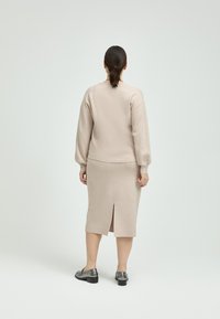 Beige Strickpullover mit Ballonärmeln, kombiniert mit einem knielangen beigen Rock mit einem Schlitz auf der Rückseite, ergänzt durch glänzende schwarze Schuhe.
