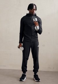 Giacca sportiva nera con zip e pantaloni abbinati, che tiene pesi silver. Le scarpe presentano accenti in bianco e nero. Tessuto liscio, design aderente.