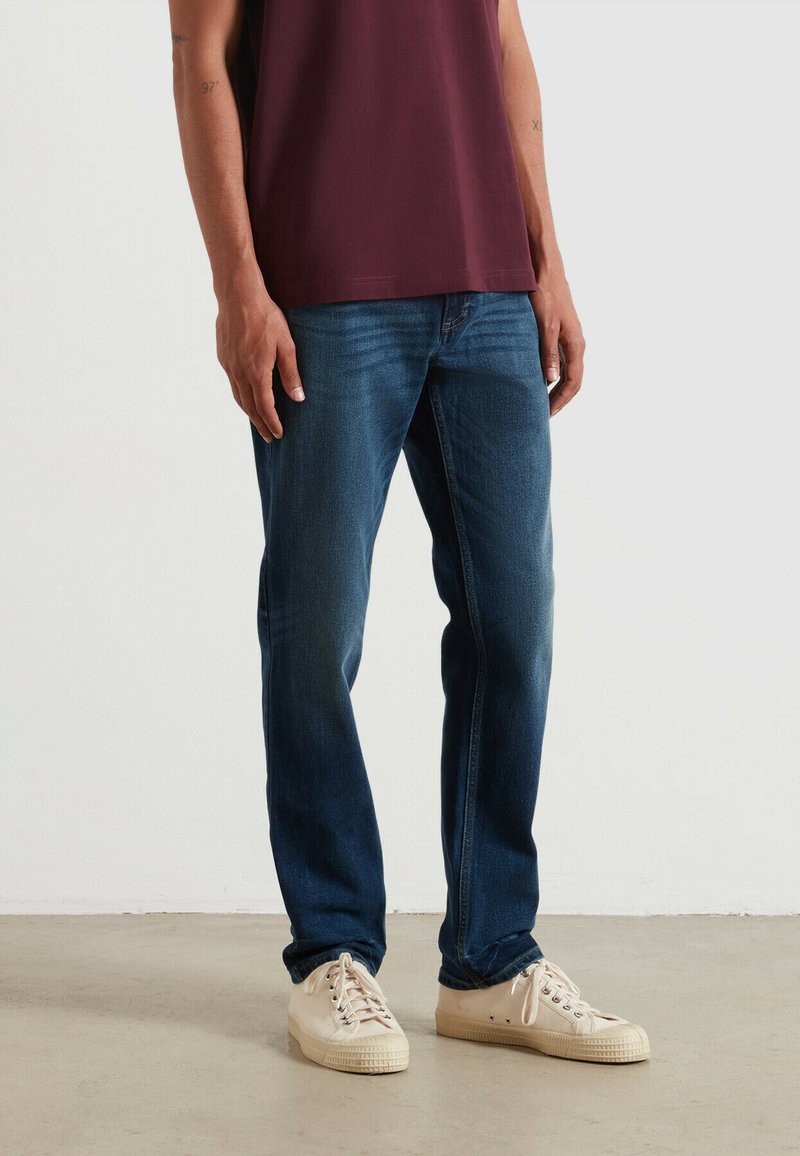 Jeans in denim blu scuro con taglio a gamba dritta, abbinati a sneakers color crema. Il materiale sembra morbido con una leggera finitura sbiadita.