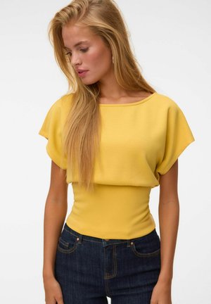 Femme aux longs cheveux blonds portant un haut jaune ample rentré dans un jean taille haute bleu foncé, regardant vers le bas à sa gauche.