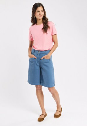 Femme portant une chemise rose à manches courtes, un short bleu mi-genoux avec des poches, et des chaussures plates marron, debout les mains dans les poches.