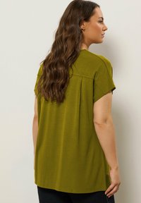 Olijfgroen kortom mouwtop met een ontspannen pasvorm, achterste plooi detail en een gladde textuur. Draagt met een donkere spijkerbroek tegen een neutrale achtergrond.
