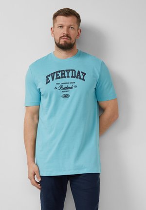 Mężczyzna z brodą w jasno niebieskim t-shircie z napisem „EVERYDAY Rethink”, stojący na tle gładkiej jasnoszarej ściany.