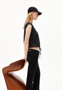 Black textured crop top with a drawstring, paired with black jeans. The model holds a smooth, curvy, brown chair. Black cap completes the look.