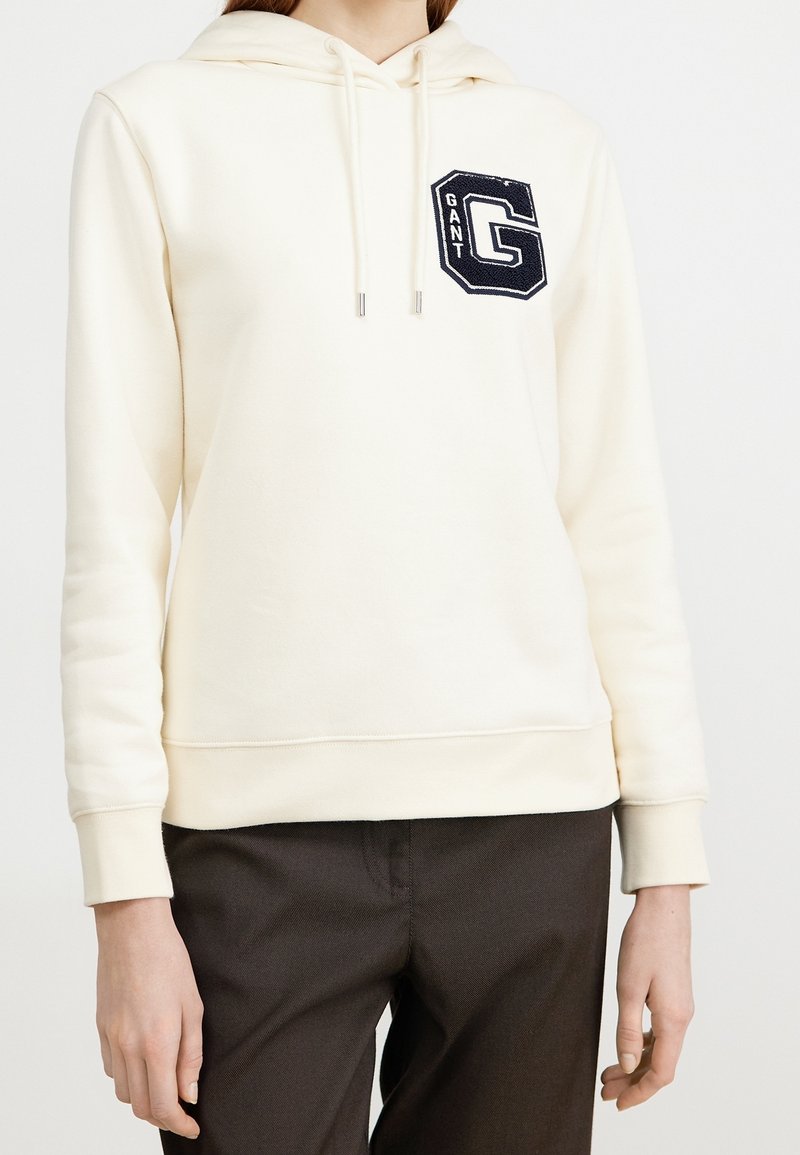 Sudadera con capucha color crema hecha de tela suave, con bolsillo frontal, cordón ajustable y detalle de logo bordado en azul marino en el pecho izquierdo.