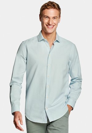 Lachende man die een lichtblauw overhemd met lange mouwen en een grijze broek draagt, staand met één hand in de zak tegen een effen achtergrond.