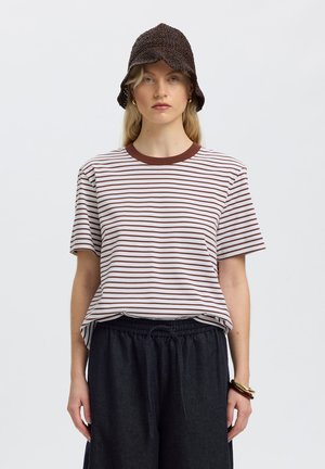 Kobieta w brązowej dzianinowej czapce typu bucket hat, białej i brązowej koszulce w paski z krótkim rękawem, luźnych czarnych spodniach oraz bransoletkach na prawym nadgarstku, stoi na białym tle.