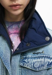 Jachetă de denim cu guler albastru, fermoar argintiu și nasturi negri. Are o emblema cu cuvântul „inspired”. Poartă peste o cămașă multicoloră.