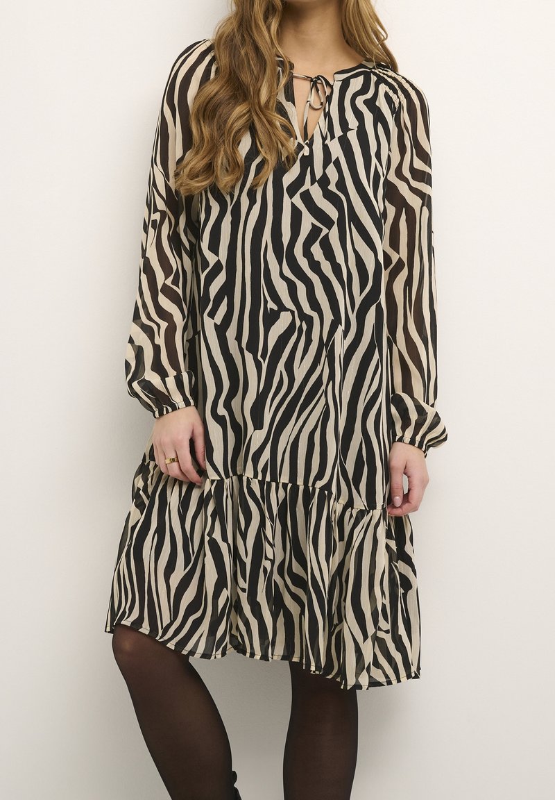 Femme portant une robe rayée zébrée noire et beige, longueur genou, manches longues, avec un ourlet froncé et un détail de lien à nouer au niveau du cou.
