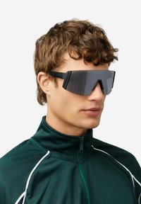 Čierne obdĺžnikové slnečné okuliare so zrkadlovými šošovkami a výrazným uhlovým dizajnom, v kombinácii s tmavozelenou bundou na zips s bielymi akcentmi.