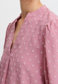 Blouse rose en tissu doux avec des motifs floraux texturés, un décolleté en V et des manches bouffantes. Détails brodés subtils et délicats sur toute la surface.