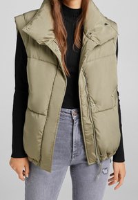Olivově zelená prošívaná vesta s vysokým límcem, zapínáním na druky a zipem. Texturovaná, prošívaná látka s bočními kapsami, nošená přes černý žebrovaný top.