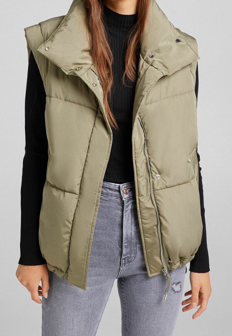 Olivově zelená prošívaná vesta s vysokým límcem, zapínáním na druky a zipem. Texturovaná, prošívaná látka s bočními kapsami, nošená přes černý žebrovaný top.
