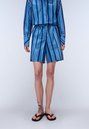 Osoba w niebiesko-białych szortach w pionowe paski oraz dopasowanej koszuli z długim rękawem, czarne klapki i tatuaż na lewej kostce.