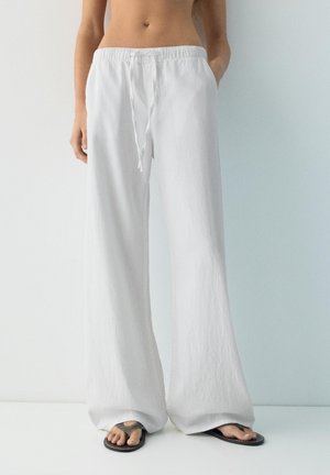 Személy laza, fehér, zsinórral állítható nadrágot és fekete papucsot visel, sima világos háttér előtt áll.