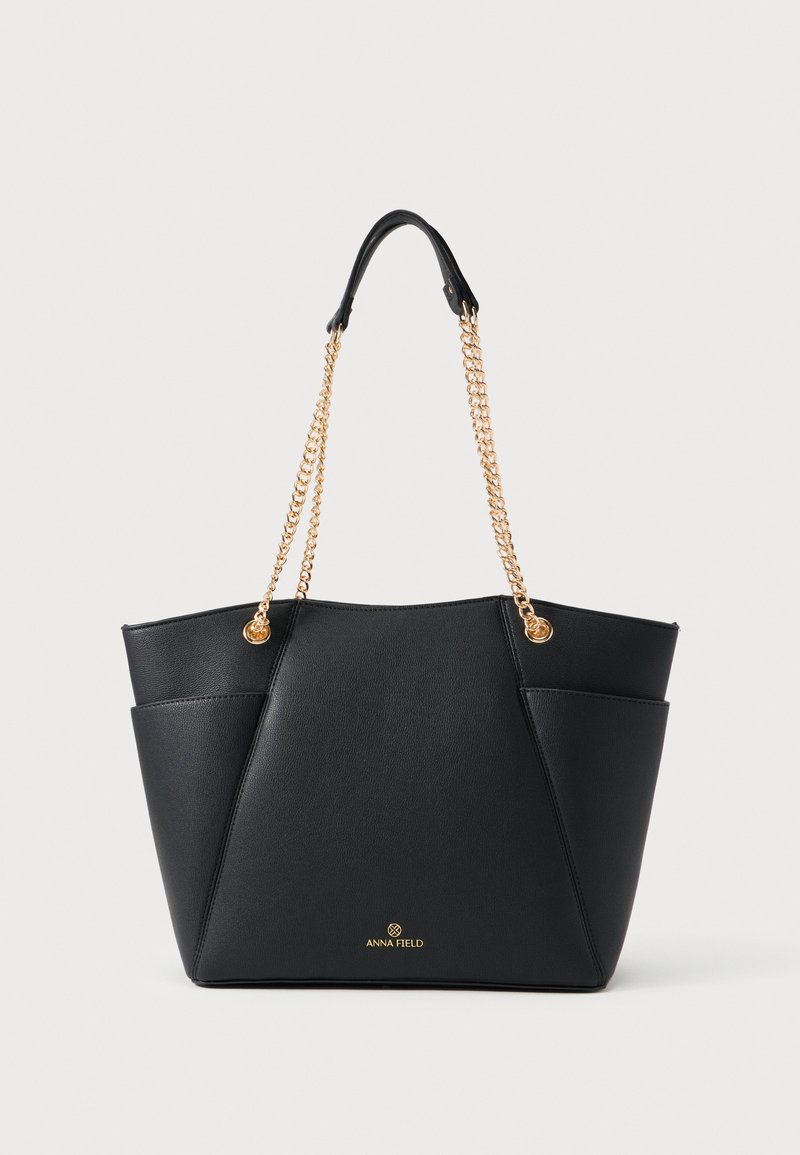 Μαύρη τσάντα tote από ψεύτικο δέρμα με γεωμετρικό σχεδιασμό, λαβές από χρυσή αλυσίδα και ανάγλυφο λογότυπο μπροστά. Λείος υφή με δομημένο σχήμα.