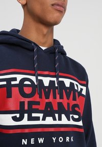 Sudadera navy con texto gráfico en rojo y blanco "TOMMY JEANS" y "NEW YORK". Cuenta con cordón ajustable con patrón a rayas. Material de mezcla de algodón.